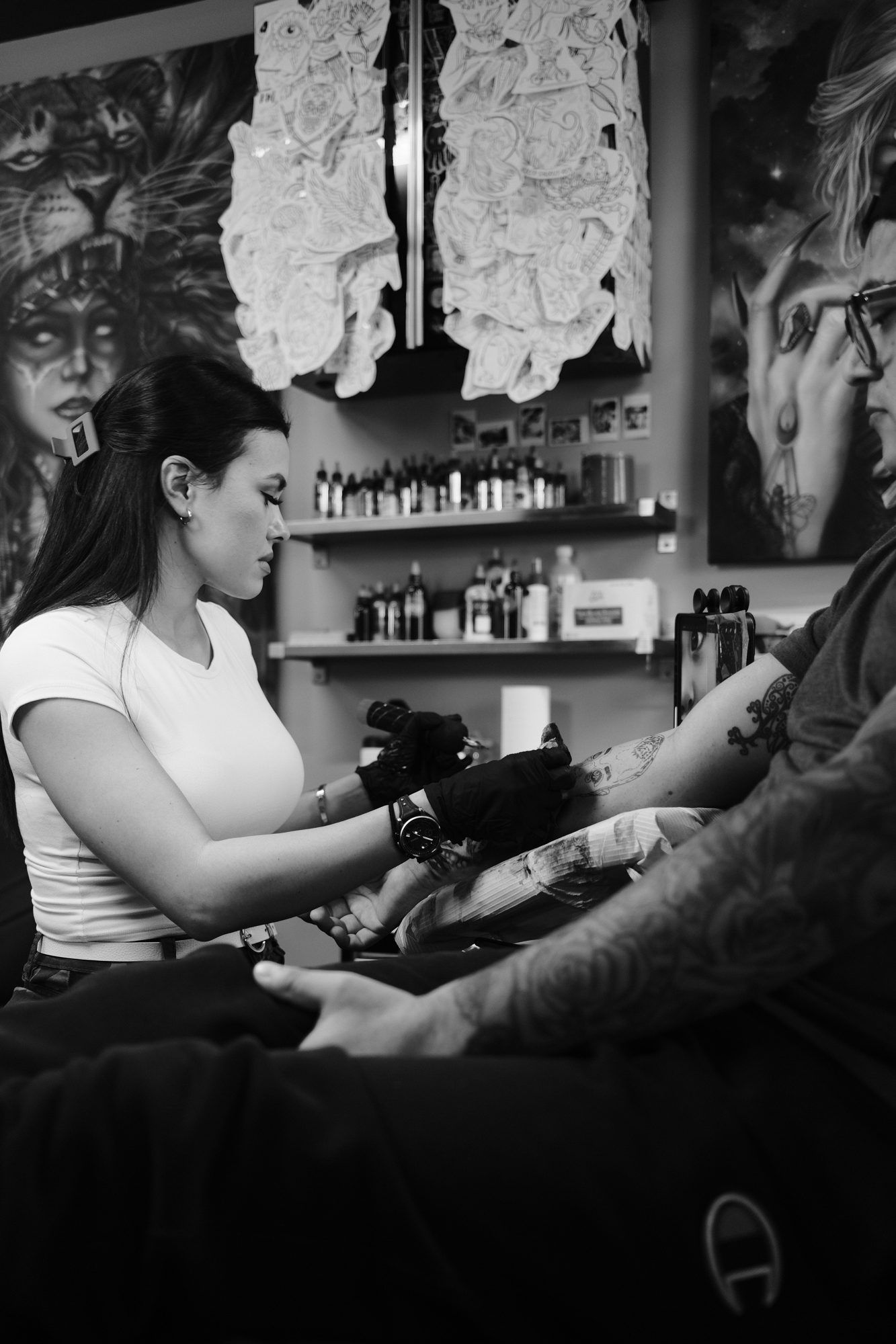 Isabella tattooing a client's forearm in the studio, with flash art and ink bottles behind her.