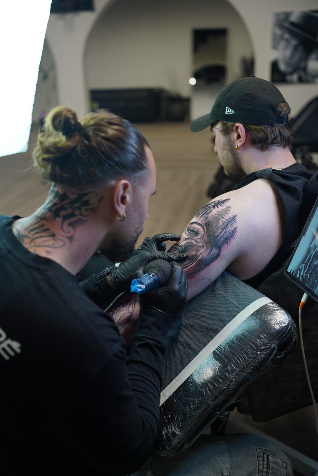 An artist tattooing a woman's portrait with pine trees on a client's upper arm in the studio.