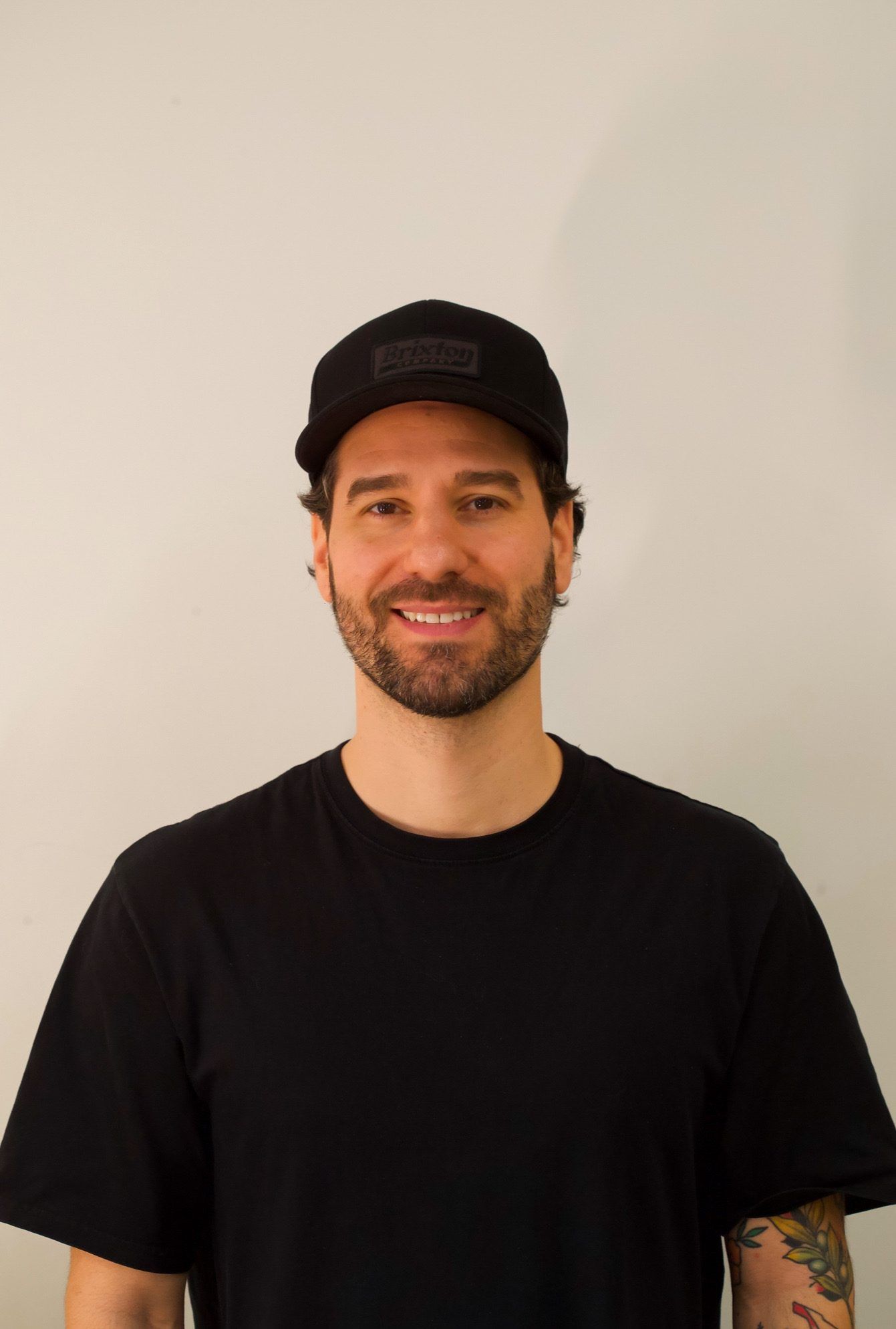 Marcos smiling in a black cap and black T-shirt against a plain wall.