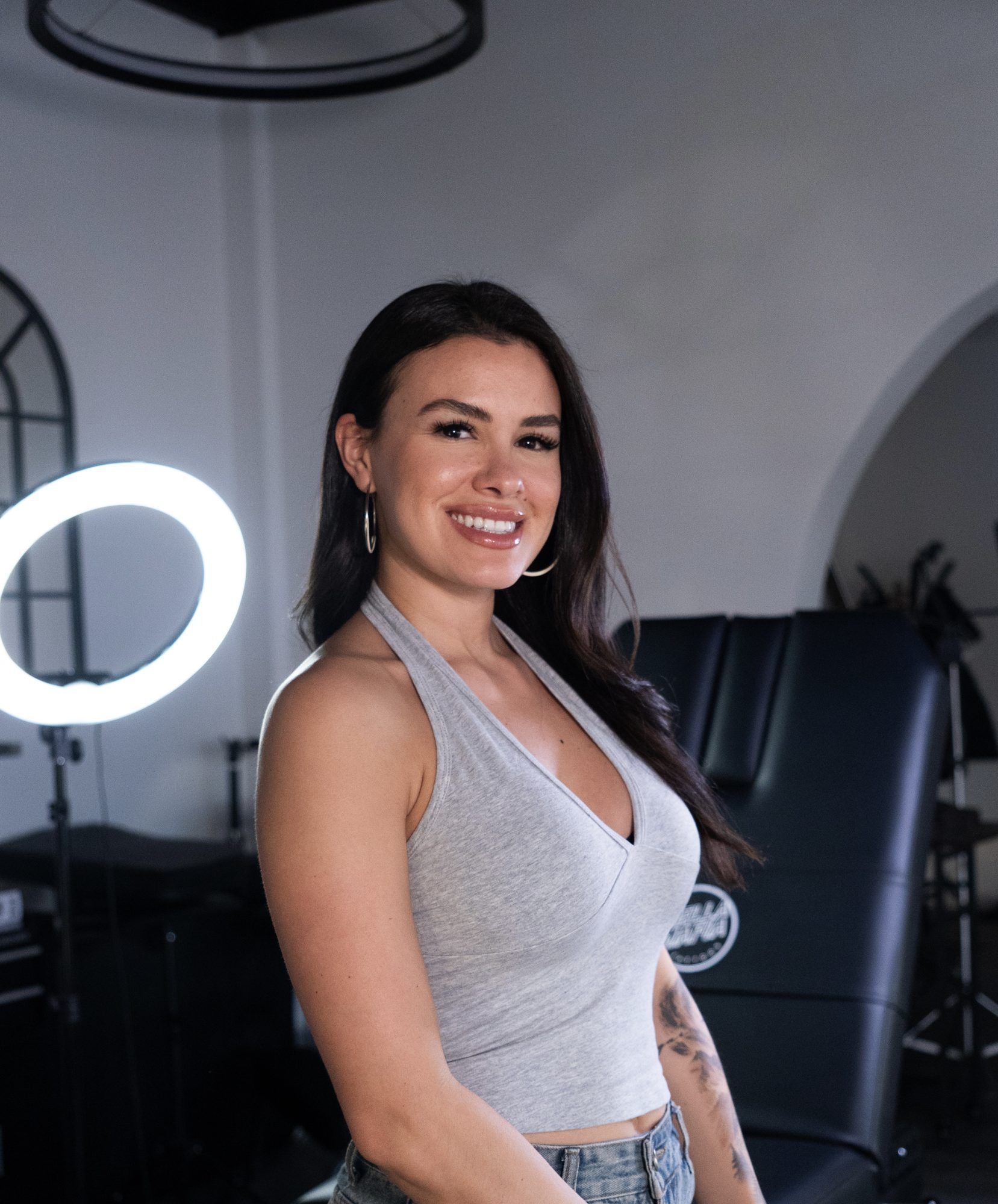 Isabella smiling in the studio beside a tattoo chair and ring light.